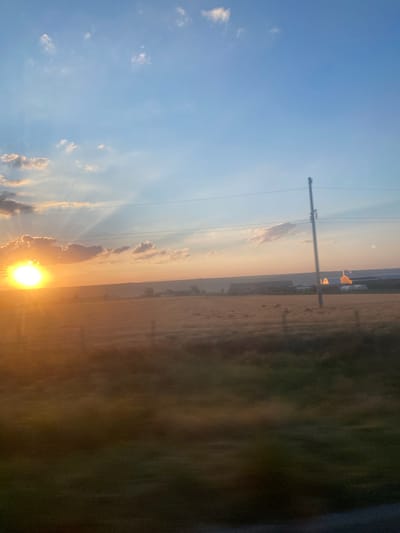 The image depicts a rural landscape at sunset, possibly in Balabanu, Moldova. The sun, a bright orb casting golden rays, is positioned low on the left horizon, partially obscured by clouds. The sky transitions from a warm orange hue near the horizon to a clear blue overhead, with scattered white clouds. In the foreground, a field of dry grass stretches towards a distant line of trees and buildings. A utility pole with power lines stands vertically in the mid-ground, slightly off-center to the right. The overall impression is one of a tranquil, late afternoon scene in the countryside.