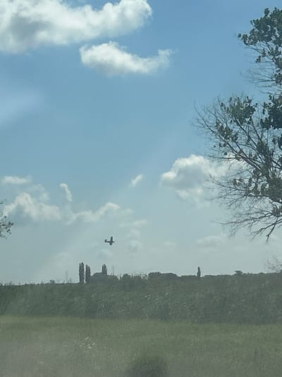 In the city of Risovyi, Russia, a plane is seen flying in the sky above a landscape. The sky is blue with scattered white clouds. A tree with bare branches is visible on the right side of the image. In the distance, there is a green, hilly terrain with some structures that appear to be buildings or trees. A person is standing on the horizon line. The foreground is a blurry green field.