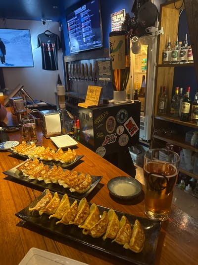 The image displays the interior of a bar or restaurant in Nozawaonsen, Japan, characterized by dim ambient lighting. A polished wooden bar counter occupies the foreground and midground. Three black rectangular plates are arranged on the counter, each holding pan-fried gyoza with crispy, golden-brown bottoms; a total of approximately 22-24 gyoza are visible. To the far right, a tall glass containing an amber liquid, consistent with beer, features a stylized 'V' logo. Adjacent to it is a small, striped ceramic dipping dish. Other items on the counter include a metal napkin dispenser, a stack of white papers, and small condiment bottles.
Behind the counter, a blue wall serves as a backdrop. A row of approximately ten beer taps with wooden handles is mounted on this wall. To the right of the taps, a black console unit adorned with various stickers (including "KEEN," "Side Show," "GOUV 500") supports a golden, cylindrical beer dispenser with a label depicting a green snowboarder. A small wooden sign on the console indicates "Wifi" with a password. Further right, a multi-tiered wooden shelf unit holds numerous bottles of spirits, including Jameson Irish Whiskey.
Above the bar, a large flat-screen television displays "WINTERLAND" and a list of items. To the left, another flat-screen television shows a snowy mountain scene with a person holding a snowboard or skis. A black T-shirt with the visible text "MORE LOVE" hangs on the blue wall to the left of the beer taps. A partial view of a person's arm and shoulder is visible at the extreme left edge of the bar counter.