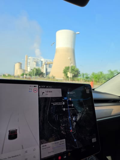 The image depicts a scene observed from inside a vehicle, looking towards an industrial power plant under a clear blue sky. A large digital display screen, characteristic of a Tesla vehicle, occupies the lower foreground.
In the background, a substantial industrial complex is visible. A prominent, light-colored cooling tower, with a white upper section and a beige base, dominates the center-right. To its left, several other industrial buildings, including a large, darker structure and a cylindrical silo, are present. Plumes of white vapor or smoke ascend into the sky from chimneys situated behind these structures. Green trees and foliage are visible along the horizon line to the right of the power plant.
The vehicle's central touchscreen displays navigational and operational information. The navigation map shows a route with a pending left turn in 1.0 km on road 554. Key locations indicated on the map include "Energoremont - Galabovo," "БРИКЕЛ" (Brikel), and "ТЕЦ Марица изток 1" (TPP Maritsa Iztok 1), confirming the geographic location as the vicinity of Galabovo, Bulgaria. The display indicates a current speed of 71 km/h, exceeding the posted speed limit of 50 km/h. Other readouts include a battery charge of 65%, the time 08:16, and an ambient temperature of 20°C. The estimated journey duration is 1 hour 42 minutes covering 108 km, with "Margi Outlet" as the destination. A stylized representation of the vehicle is shown on the left side of the screen, moving along a digitized road. The vehicle's dashboard is visible below the screen, and a portion of the windshield shows reflections of the sky. A small, dark, elongated object is present on the windshield, positioned against the blue sky to the right of the main cooling tower. The sky is bright blue with some diffused white vapor from the power plant.