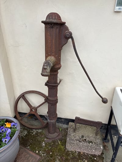 An exterior shot shows a vintage, rusty, cast-iron water pump mounted horizontally on a pale, off-white wall. The pump has a lever with a spherical end, a spigot, and a wheel. Next to the pump, on the ground, is a mossy area with a concrete block, and a planter containing purple and yellow flowers. Above the pump, the corner of a white structure is visible. The scene appears to be located outdoors, likely in the city of Minsterley, United Kingdom.
