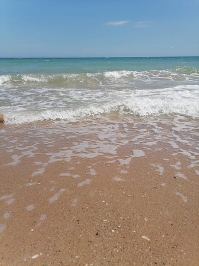 The image depicts a low-angle view of a beach in Durankulak, Bulgaria. The foreground is dominated by wet, sandy beach with small shells and pebbles. Gentle waves with foam are washing ashore, covering the sand in a thin layer of water. The middle ground features the breaking waves of the sea, which appear clear and a greenish-blue color. The horizon line is distinct, separating the sea from a clear, light blue sky with a few wispy clouds. No people or man-made objects are visible in the frame, emphasizing a natural, serene beach environment.
