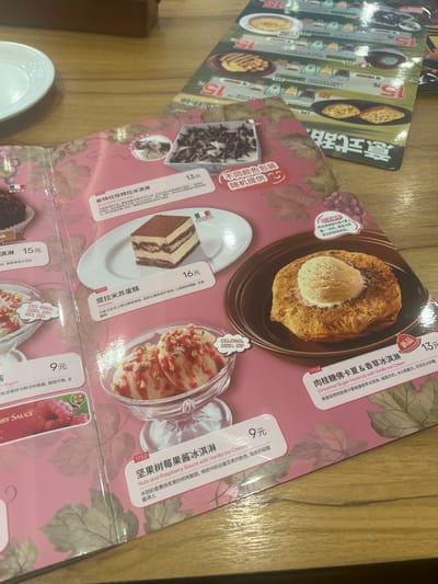 This is a top-down, close-up shot of a menu or flyer laid out on a wooden table. The menu features vibrant pink and green designs with illustrations and photographs of desserts. Several food items are visible and described with Chinese and English text.
To the left, a small white bowl with a white dessert and red sauce is partially visible. Below it, a menu item with the heading "Berry Sauce" is shown with a close-up of raspberries.
In the center, a slice of tiramisu is displayed on a white plate, described as "Tiramisu" with a price of 16 Yuan. Below this, a dessert in a glass bowl with two scoops of ice cream, drizzled with red sauce and topped with chopped nuts and seeds, is shown. This item is labeled "Nuts and Raspberry Sauce with Vanilla ice Cream" with a price of 9 Yuan.
To the right, a circular baked item topped with a scoop of vanilla ice cream is presented on a brown plate. This is described as "Cinnamon Sugar Focaccia with Vanilla Ice Cream" with a price of 13 Yuan. A small circular image shows a scoop of ice cream.
The menu also includes other dessert images and descriptions, with prices ranging from 15 Yuan upwards. The overall impression is of a dessert menu from a cafe or restaurant. The context of Baiyun District, China is not directly inferable from the image itself, but could be assumed based on the language used and the typical offerings in such establishments in China.