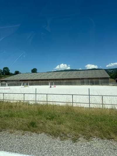 A long, wooden building with a grey, patterned roof stands behind a fenced, white arena. The building has the words "CENTRE EQUESTRE" and "JURA SUD" written on its facade, along with a phone number. Several horse jumps are set up in the arena. The area is surrounded by grass and trees, with a blue sky and clouds above. The scene appears to be in Charchilla, France.