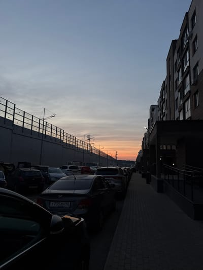 The image depicts a street scene at dusk in the city of Военнослужащих Кп, Russia. On the right side of the frame, a modern multi-story apartment building with numerous windows and brick accents lines the sidewalk. The sidewalk itself is paved with dark rectangular bricks. A row of parked cars stretches along the left side of the street, with a dark-colored sedan in the foreground and a darker vehicle with a license plate reading "T 735 AM 40" visible behind it. Further down the street, more cars are parked. Beyond the cars on the left, a high concrete wall with a metal railing and decorative elements runs along the edge of the street. Electrical poles and wires are visible in the distance, silhouetted against a sky with a gradient of pale blue and soft orange hues, indicating the setting sun. The overall scene is one of urban infrastructure under the fading light of day.
