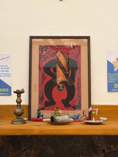The image displays an indoor scene featuring a framed piece of art, various decorative objects on a wooden shelf, and two printed notices on a light-colored wall.
Centrally placed on the light, textured wall is a dark-brown framed artwork. The artwork itself is a striking graphic print, possibly a woodcut or linocut, depicting two stylized, solid black figures against a vivid red background patterned with black textures. One larger figure, which appears to be a woman with distinct hair or a headdress, is shown embracing or holding a smaller, infant-like figure. The overall impression of the art is one of connection or nurturing. Reflections are visible on the glass covering the print, suggesting a light source in the room.
To the right of the framed art, a wooden shelf or ledge holds an assortment of small decorative items. These include a colorful mermaid figurine with a blue tail, a bundle of white material (possibly sage or palo santo) tied with string, a small green succulent-like plant, and a black figurine that resembles an animal. Further to the right on the shelf, a brass or bronze candlestick holder with a melted purple candle stub is visible, along with other small, indistinct objects like stones or shells. The wooden shelf itself has a light, natural finish.
The wall on which the art and notices are displayed is a uniform light cream or off-white color with a subtle texture. To the far right of the wooden shelf, a section of a rough, dark stone wall is visible, providing a contrasting background texture.
Two printed notices are affixed to the light wall. One, partially visible above the framed art, is on a blue background and contains text such as "Ven y celebra," "bailando a la Virgen de la Candelaria," "DJ set de," and "Iturbe." Another partially visible notice, also on a blue background, is located below the framed art and includes phrases like "ente eurore," "Sábado de enero," "7:00 PM," "tamales y," "gos amargos," and "Michoacan." These notices appear to be advertisements or announcements for local events.