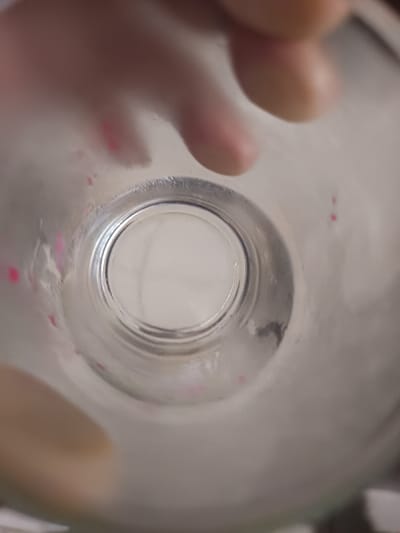 The image displays a direct, top-down, interior view of a transparent, cylindrical vessel, likely made of glass or clear plastic. The vessel's interior surface exhibits multiple small, irregularly shaped pink-red smudges or residue particles, primarily visible on the left and right inner walls. The circular base of the vessel is visible at the bottom center, appearing light-colored and out of focus. The top portion of the frame shows blurred human fingers, suggesting a hand is holding or otherwise interacting with the vessel from above. The vessel appears largely empty. No specific details regarding the broader environment or a precise location within the city of Ouderkerk aan de Amstel, Netherlands, are discernible from this image.