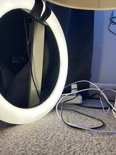 The image shows a close-up view of a ring light resting on a surface, possibly a carpeted floor. The ring light is white with black tape on one side. Several cables are visible, including a black power cord and some clear-covered cables. A white power adapter with multiple outlets is plugged in. The background includes a dark gray fabric and a wall painted in a shade of blue or gray. There is no information in the image that suggests its location is in the city of Raduga, Russia, or any specific location. The scene appears to be an indoor setting.
