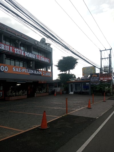 The image shows the exterior of "Sturgis 69" Boutique Hotel, which includes a grill and bakery. A large banner advertises "137K PORSI 2-3 ORANG" and "SAE-PULEN-RAOS 2.600". Next door is "Sturgis Bakery" and "HOTEL SHABU - SHABU & GRILL". The parking lot is paved with light-colored blocks and marked with orange cones and posts. Overhead, a tangle of power lines crisscrosses the sky.