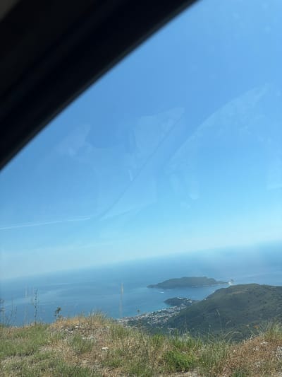 The image shows a scenic overlook of the Adriatic Sea, with a view of the coastline and islands near Stanišići, Montenegro. In the foreground, dry grasses and vegetation cover a hillside, suggesting a natural, undeveloped area. The middle ground features the blue expanse of the sea, dotted with a few small boats. On the right side of the image, a coastal town with buildings can be seen nestled between green hills and the sea. To the left of the town, a larger, lush green island is prominent in the water. The sky above is clear and blue, indicating a bright, sunny day. Reflections are visible on what appears to be a window, possibly from a vehicle, with a dark frame along the top left edge.