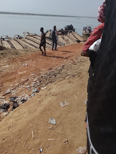 The scene depicts a riverine ferry landing in Ibi, Nigeria, under a hazy, light-colored sky. The foreground features a reddish-brown dirt embankment, heavily littered with numerous plastic bags, clear plastic wrappers, and scattered debris. Partially visible on the right, in the immediate foreground, is the right side of an individual wearing dark trousers and a red-patterned shirt, with a patterned head covering.
In the midground, a sloping dirt path leads down to the water. Two male subjects are present: one dark-skinned male in a dark t-shirt and dark shorts walks barefoot towards the right across the dirt path, with his left arm slightly raised. Behind and to his right, a second dark-skinned male, dressed in a blue long-sleeved shirt and dark trousers, stands facing away from the camera, looking towards the river.
The riverbank itself is lined with multiple wooden, flat-bottomed ferries or barges, most of which appear to be docked or beached. These vessels have slatted wooden decks. One ferry, visible in the mid-left, has a dark-colored vehicle, possibly a truck or utility vehicle, loaded onto its deck along with other indistinct equipment. Further out on the river, to the mid-right, another ferry is positioned in the water, appearing to be actively loaded with multiple people and at least one vehicle. The river itself is wide and calm, with a distant treeline visible on the far bank. A small, indistinct boat is also visible further out on the water.