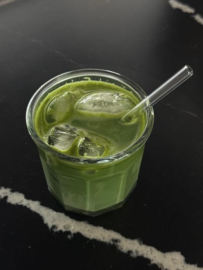 An overhead shot depicts a glass of green liquid, likely matcha, filled with ice and a clear straw, placed on a black countertop with a marbled white vein. The location is potentially in Zapopan, Mexico, although there is no explicit evidence of this in the image.