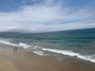 The image depicts a wide-angle view of Hermosa Beach, California, United States of America. The scene is dominated by the ocean and the sandy shore. The ocean is a deep blue-green, with visible waves breaking near the shore. The sky above is a mix of blue and white clouds.
On the wet sand of the beach, two individuals are walking. One person appears to be wearing a pink top and dark shorts, while the other is in darker clothing. They are walking in the same direction, away from the water. Further out in the water, two individuals are visible. One appears to be body surfing or paddling on a surfboard, and the other is closer to the shore, possibly also engaged in a water sport.
In the background, the coastline of Hermosa Beach is visible, with buildings and hills lining the shore. A pier extends out into the ocean from the left side of the frame. The sand of the beach is a light brown color, wet and reflective near the water's edge. The overall impression is a typical summer day at the beach, with people enjoying the ocean and the shoreline.