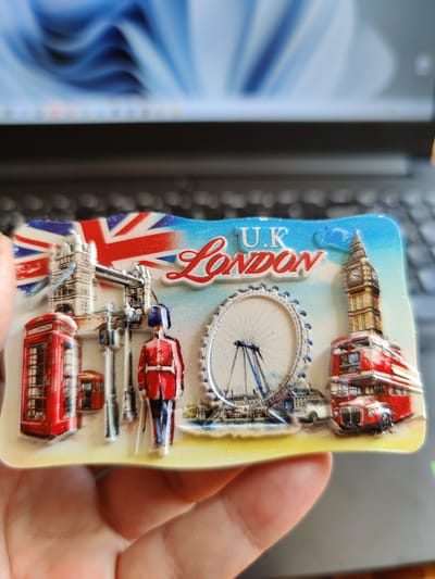 A hand holds a souvenir magnet depicting London landmarks. The magnet features the Union Jack flag at the top, followed by the Tower Bridge, a red telephone booth, a Beefeater guard, the London Eye, and Big Ben with a double-decker bus in front of it. The background shows a blurred image of a laptop screen. The location in Rybnitsa, Moldova is not determinable from the image.