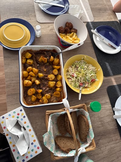 The image depicts a table laden with food, possibly in Bruchköbel, Germany, though no specific landmarks are visible. A rectangular baking dish filled with roasted meat and potatoes sits centrally. A yellow bowl contains a salad, and another white bowl holds meatballs and French fries. Bread is presented in a wicker basket lined with a floral cloth. Blue and white plates, cutlery, and a tissue box with colorful polka dots are also visible on the wooden table. No people are discernible in the image.