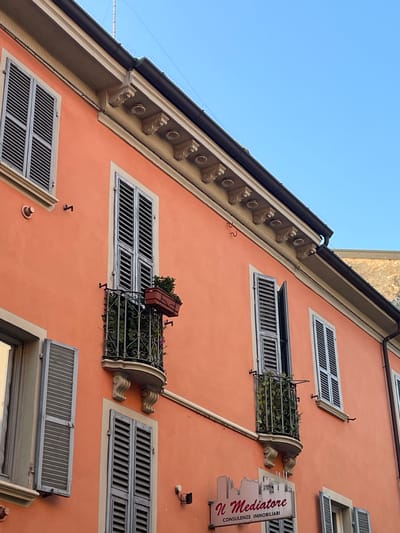 The image shows a close-up view of an orange building facade in Piacenza, Italy. The building features several windows with closed, grey shutters. Two balconies with ornate metal railings and greenery are visible. One balcony has a planter box with a plant. A sign for "Il Mediatore Consulenze Immobiliari" is mounted on the lower part of the building. The sky is clear and blue. The architectural style suggests an older building, typical of Italian city centers.