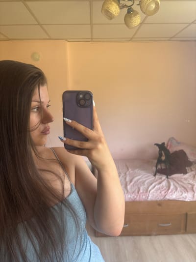 The media file shows a young woman taking a mirror selfie in what appears to be a bedroom. The woman has long, dark brown hair and is wearing a light blue, glittery dress. Her nails are painted with a white and blue design. She is holding a dark purple smartphone in her right hand, angled to capture her reflection.
The setting includes a light-colored wall and a tiled wall with vertical lines. In the background, reflected in the mirror, is a bed with pink and white bedding, and a wooden dresser or cabinet with handles. A light fixture with multiple globes is visible on the left side of the frame. The overall lighting suggests it might be daytime or a well-lit interior. The woman's expression is neutral, with her gaze directed towards the phone. No visible text is present in the media.