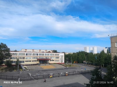 This is a medium-angle, eye-level shot of a school courtyard on a clear day, with a prominent school building in the midground. The building, likely a school, has a light beige facade with multiple rows of windows and a central entrance with wide stairs leading up. Adjacent to the entrance is a large, colorful mural. The courtyard is paved and features a black metal fence separating it from a sidewalk and street area. Several trees line the perimeter of the school grounds, and in the background, modern residential buildings, including tall white towers, are visible. A security camera is mounted on a pole to the right of the school building. The sky is a vibrant blue with scattered white clouds. The timestamp "21.07.2026 16:43" is visible in the bottom right corner, and "POCO X6 Pro 5G" is in the bottom left. The scene is located in Kogalym, Russia.