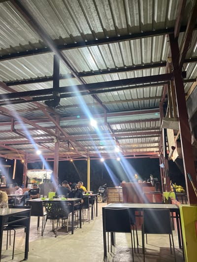 An outdoor restaurant at night, illuminated by bright lights along a metal-framed roof. People are seated at several tables. A motorcycle is parked near a wooden structure. A metal roof and support beams make up the ceiling, with a hanging fan visible. The floor is concrete.