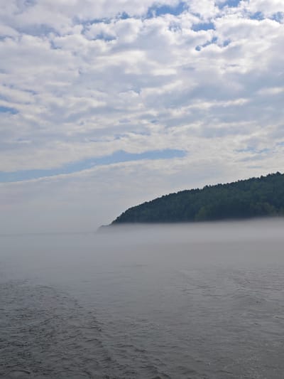 The image depicts a body of water shrouded in a thick layer of fog, obscuring the horizon and a distant, tree-covered hillside. The water in the foreground shows ripples and wake, suggesting movement. The sky above is partly cloudy, with patches of blue sky visible between white clouds. The overall scene conveys a sense of atmosphere and natural beauty. The location is the Yenisei River near the village of Atamanovo, Krasnoyarsk Krai, Russia.