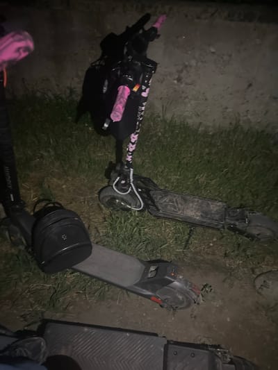 The image depicts a nighttime outdoor scene in Tazha, Bulgaria, featuring four electric scooters positioned on a surface of mixed grass and bare earth, adjacent to a textured concrete or stone wall in the background.
In the center-right, a black Kugoo electric scooter with distinct pink grips and pink cherry blossom graphics on its stem stands upright but slightly leaning. A small, black cylindrical storage bag is attached to its handlebar stem. The scooter shows signs of dirt on its wheels and lower chassis.
To the left, a portion of a black Xiaomi electric scooter is visible, also with a pink grip and a similar black cylindrical storage bag resting on its deck. Its stem features the "Xiaomi" branding.
In the foreground, a third black electric scooter's deck is visible, ribbed for grip. A person's right foot, clad in a dark sneaker, is positioned near this scooter's deck.
Horizontally across the right side of the frame, a fourth black electric scooter lies flat on the ground, appearing dirty and well-used. Its deck and wheels are covered in what appears to be dried mud or dirt.
The ground consists of patches of dark green grass, drier brown grass, and exposed dirt. The lighting is dim, suggesting night conditions, with an unseen artificial light source illuminating the scene, creating soft shadows.