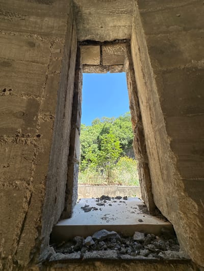 The image shows a view through a rectangular opening in a concrete structure. The structure's walls are rough and weathered, with visible aggregate in the concrete. Through the opening, a patch of bright blue sky is visible above green trees and vegetation. A chain-link fence can also be seen. The scene is likely located in Donji Katun, Montenegro. The focus is on the opening and the view beyond.