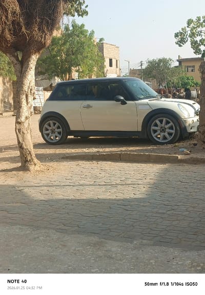 A white Mini Cooper with a black roof is parked on a sandy street. The car faces right, with its front near a rough-hewn curb. A gnarled tree trunk dominates the left foreground, casting a shadow on the stone-paved street. In the background, buildings, trees, and people can be seen. The image is taken in daylight.