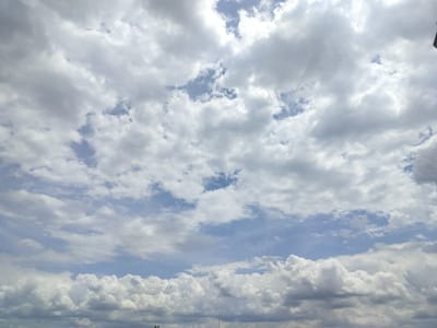 A wide shot captures a cloudy sky above Gorno Sahrane, Bulgaria. Puffy white and grey clouds dominate the frame, with patches of blue sky visible between them. The lower edge of the image shows a sliver of a cityscape, with dark outlines of buildings and structures suggesting an urban environment. The lighting indicates it is daytime, likely overcast.