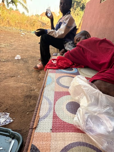 The scene is located in Oturkpo, Nigeria. In the foreground, an adult male is lying on his right side, appearing to sleep or rest on a mattress with a multi-colored geometric pattern. He wears a red garment. A crumpled red fabric item lies near his head, and a translucent plastic bag rests by his legs. To the left, a dark rectangular tray contains a dark liquid and small white fragments on the reddish-brown dirt ground. In the mid-ground, a child or young person sits, looking directly towards the camera, holding a crumpled clear plastic bottle in their right hand. This individual wears a blue and white long-sleeved jacket, dark trousers, and light-colored sandals. Another person's leg is partially visible behind the child. The background features dry, tall brown vegetation and a distant building structure under natural daylight. A reddish-brown building wall is visible on the right. The dirt ground contains scattered white plastic debris.