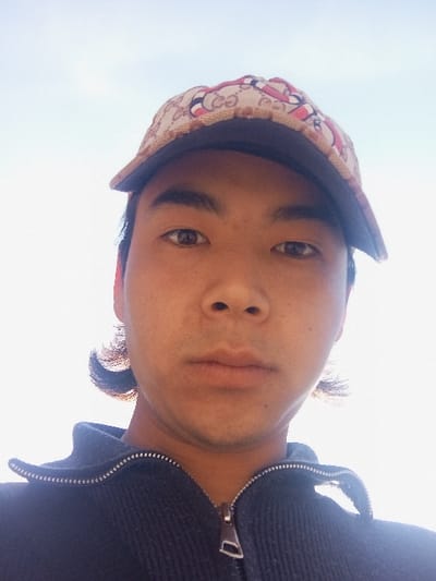 A low-angle, close-up portrait depicts a single male subject, appearing to be of East Asian or Central Asian ethnicity, estimated as a young adult. He is positioned looking directly forward. The subject wears a beige baseball cap featuring a repeating "GG" pattern and a red and black serpent design, with a dark brim. Below the cap, dark hair is visible extending slightly past the ears. His attire includes a dark-colored, likely navy blue or black, ribbed knit jacket or sweater with a half-zip closure and a silver-toned zipper pull. Subtle facial hair is present on his upper lip and chin. The background is an overexposed, bright light-blue sky, indicative of an outdoor environment with direct overhead illumination. The scene is located in Takhtakuprik, Uzbekistan; however, no specific visual elements within the image directly identify this city or a particular point within it.