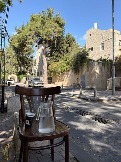 The image depicts an outdoor street scene in West Jerusalem, Israel, under a clear blue sky. In the foreground, a dark wooden chair is positioned on a paved stone sidewalk. On the chair's seat rests a clear glass pitcher containing water, exhibiting condensation, next to a black disposable cup with white stripes and a red heart graphic. A black metal table base is partially visible to the left.

The midground features a paved street with a linear storm drain grate, showing dappled sunlight and shadows. A silver SUV is observed driving away from the viewer on the street. Multiple grey bollards line the street's edge, bordering a stone retaining wall partially covered in green foliage. Above the wall, a multi-story building constructed of light-colored stone is visible, featuring several rectangular windows, a distinctive round window, and a white vent on its roof. Utility poles with overhead wires are present on the left and right.

The background is dominated by numerous tall green trees, primarily pine trees, extending above the buildings, and a clear blue sky. A stone archway structure is visible in the distance along the curving street.