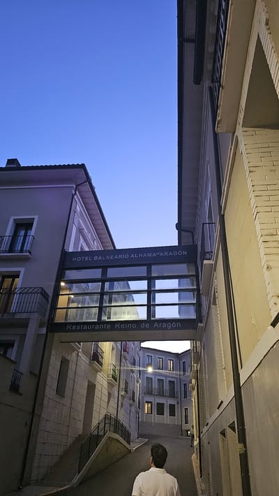 The image captures a nighttime scene in a narrow street in Alhama de Aragón, Spain. The sky is a deep, clear blue, indicating it is twilight or early night. Buildings line both sides of the street, with lights on in some of the windows. The architecture suggests an older European town.
In the foreground, a building features a prominent sign with text that appears to be in Spanish. The sign is mounted above a glass-fronted entrance. The text on the sign reads "HOTEL BALNEARIO ALHAMA DE ARAGON" vertically on the left side, and "Restaurante Reino de Aragón" vertically on the right side. This indicates the presence of a hotel and a restaurant.
To the right of the frame, a person wearing a white shirt is seen from behind, walking away from the camera down the street. The person is partially illuminated by the streetlights. The street itself is paved, and there are railings and balconies visible on the buildings. The overall atmosphere is quiet and serene, typical of a Spanish town in the evening.