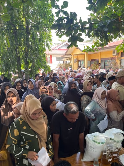 In the city of Pulodang, Indonesia, a crowd of people, predominantly women wearing hijabs of various colors and patterns, are gathered outdoors. They stand in a line, with many looking towards the right side of the frame. The background features a yellow building with a red-tiled roof, suggesting it might be a public facility. Large trees with green leaves hang over the scene, providing shade. In the foreground, two large white sacks with text visible and two clear plastic bottles containing a brown liquid are placed on a wooden table. A man in a black t-shirt is looking down at the table. The overall atmosphere appears to be one of organized distribution or waiting.