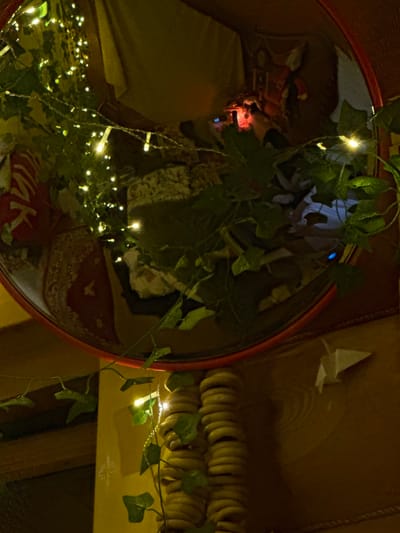 A red-framed circular mirror reflects a scene of draped fabric, fairy lights, and a plant with green leaves. A small origami paper crane is visible near the mirror’s edge. To the right, a row of stacked bagels or round pastries hangs on a yellow surface with green ivy. The lighting is dim and warm, creating a cozy, cluttered aesthetic.
