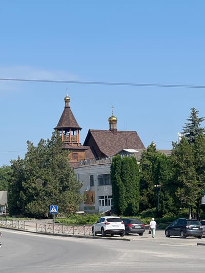 The image depicts a view of a street scene in Chemerivtsi, Ukraine, with a church prominently featured. The church has a tall, wooden bell tower topped with a golden dome and cross. Adjacent to the bell tower is the main church building, also with a brown tiled roof and a smaller golden dome. Several mature evergreen trees and bushes are situated in front of the church. A light-colored building with a mural on its side is visible behind the trees. In the foreground, a street with parked cars is visible. A white SUV, a dark hatchback, and a dark sedan are parked on the side of the road. A pedestrian walkway is marked with a blue and white sign featuring a pedestrian icon. A woman dressed in a white top and light-colored pants is walking away from the camera towards the parked cars. The sky is clear and blue.