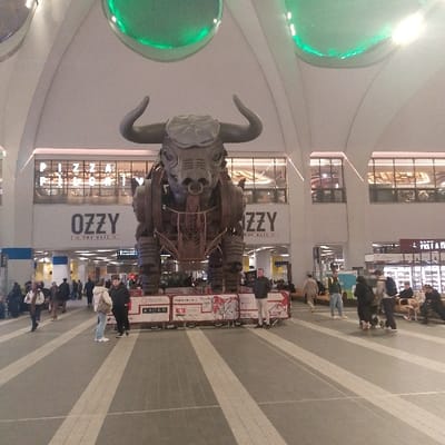 The image depicts the Grand Central concourse at Birmingham New Street Station in Birmingham, United Kingdom. A prominent, large, grey, robotic-style bull sculpture, identified by adjacent signage as "Ozzy The Bull," stands centrally on a low, banner-decorated platform. The bull features large horns, a nose ring, and visible mechanical components.
The concourse has a high, arched white ceiling with multiple circular green light fixtures. The floor is tiled with alternating light and dark grey stripes. In the background, an upper level features various storefronts, including "Pizza Pilgrims" visible above the "Ozzy The Bull" sign, and "Pret A Manger" further to the right.
Numerous people are present throughout the scene. Individuals and small groups are walking, standing, or sitting. A man in a dark jacket and light trousers stands near the base of the bull on the right, facing towards the viewer's left. Two individuals are conversing near the front left base of the bull. Others are dispersed across the concourse, appearing to be commuters or shoppers. Their clothing suggests cool weather conditions.