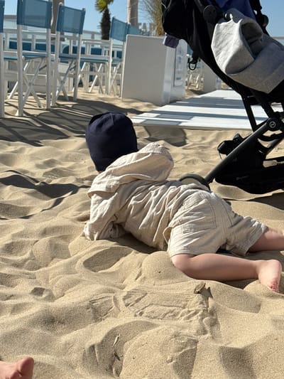 The image depicts a young child crawling on a sandy beach in Middelkerke, Belgium. The child, viewed from behind, is on all fours, clad in light-colored, possibly linen, shorts and a long-sleeved top with a hood, along with a dark navy cap. Their face is not visible.
To the left of the child, a black baby stroller or buggy, bearing the visible "Cybex" brand name on its frame, is partially captured. A grey fabric item is placed within the stroller. In the bottom right corner, a portion of an adult's bare foot with painted toenails is visible.
The scene is dominated by vast amounts of light-colored sand, showing textures and shadows indicative of bright sunlight. In the background, rows of white-framed beach chairs with blue fabric are arranged, suggesting a beach club or resort setting. A palm tree is also faintly visible on the far left. The clear, sunny weather casts distinct shadows across the sand. A small, dark, twig-like object is partially embedded in the sand near the child. The overall atmosphere suggests a relaxed outdoor activity on a bright day.