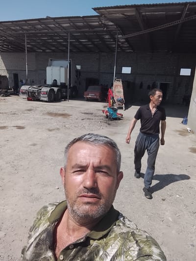 The image depicts an outdoor industrial or workshop yard in Sabir Rakhimov, Uzbekistan, under bright daylight. In the foreground, a middle-aged man with graying hair and a beard, wearing a green and white patterned polo shirt, is taking a selfie, looking directly at the camera. His face appears slightly shiny, suggesting warmth.
In the mid-ground, a man of Asian ethnicity, dressed in a dark polo shirt and dark loose-fitting pants, is walking from left to right, looking towards his right. He casts a distinct shadow on the dusty, gravelly ground.
Behind the men, the yard contains several vehicles and structures. A large white semi-truck tractor unit is parked beneath an open-sided shelter with a corrugated metal roof supported by metal beams. To its right, a red vintage-style car, possibly a Lada, is also parked under the shelter. The shelter extends over a building constructed from light-colored concrete blocks, which has multiple doorways and two rectangular, light-colored openings that may be windows or vents. On the far left, another red vehicle is partially visible.
In the center of the background, a red and blue engine hoist with a blue engine block attached to it suggests ongoing mechanical work. A large blue plastic water bottle is visible on the right side of the frame. The sky above is clear and blue. The strong shadows cast by objects and people indicate that it is likely midday or early afternoon. Faint text "DWOM" is visible on the mudflaps of the semi-truck.