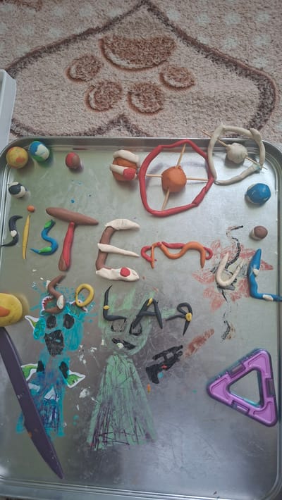 This image displays a metal tray filled with various objects, predominantly made of playdough. The playdough has been shaped into letters spelling "SISTER SOUL" and also into several small figures, some resembling planets or celestial bodies, with toothpicks inserted into them. There are also some crudely drawn figures in green and blue on the tray. A purple plastic knife lies on the left side of the tray. A magnetic building toy in the shape of a triangle is positioned in the bottom right corner. The tray rests on a patterned rug with a paw print design. The scene is likely indoors, on a flat surface, possibly a table. The location in Voinova, Moldova, cannot be determined from the visual information provided.