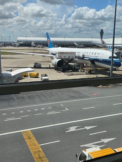 This is an eye-level, outdoor, medium shot of an airport tarmac on a partly cloudy day. In the foreground, a dark grey asphalt road has white lane markings and directional arrows, as well as a yellow painted instruction in Chinese characters. In the mid-ground, a large China Southern Airlines passenger jet is parked at a gate, connected to the terminal by a jet bridge. Luggage carts and service vehicles are near the aircraft. The terminal building in the background has a distinctive, modern, silver, honeycomb-like facade. A control tower is visible in the distance to the right of the terminal.