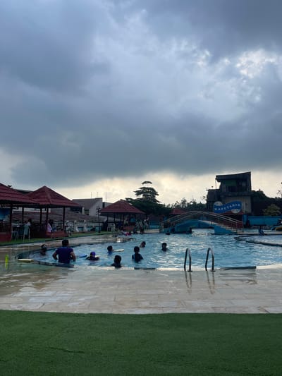 The image shows an outdoor swimming pool scene in Ngasinan, Indonesia. The sky is mostly overcast with heavy, dark clouds. A swimming pool is visible, occupied by several individuals swimming or standing in the water. There are pool ladders visible on the side. A tiled area surrounds the pool, with a section of green artificial turf visible on one side. Several gazebo-like structures with red roofs are adjacent to the pool. In the background, there is a building with a sign that says "Kafetaria" [sic]. Other buildings and vegetation are present in the surrounding area.