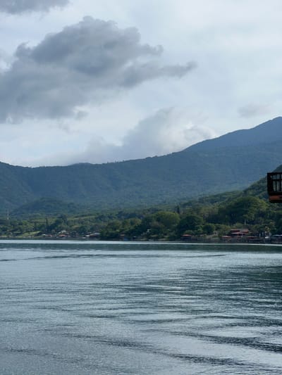 The image displays a wide view of a large body of water, likely a lake, bordered by mountainous terrain under a cloudy sky. The scene is situated in the city of Unknown, El Salvador. The sky is predominantly overcast, featuring dense grey clouds with varying degrees of brightness. The background is dominated by a series of steep mountains, which are heavily covered in lush, dark green foliage, indicative of a forested environment. The upper parts of these mountains are partially shrouded in mist or lower-lying clouds.

Along the base of the mountains, at the water's edge, a series of human-made structures are visible. These appear to be buildings, possibly homes or resorts, characterized by reddish-brown roofs. Some of these structures are built directly on the shore, while others extend over the water on what appear to be stilts or piers. On the left side of the distant shoreline, a tall, slender, dark-colored mast or tower, resembling a cellular antenna, is prominently visible rising above the tree line.

The foreground and mid-ground are occupied by the surface of the water, which exhibits numerous parallel ripples and waves, suggesting gentle movement or a breeze. The water's color is a calm, desaturated blue-grey. In the upper right corner of the frame, a segment of a dark wooden structure, appearing to be a balcony or an elevated deck with a railing, extends into the scene. There are no individuals or overt activities discernible within the frame.