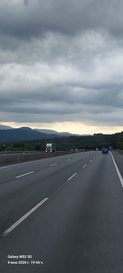 The image shows a perspective view of a highway in Maçanet de la Selva, Spain, under a cloudy sky. The road has multiple lanes marked with white lines. In the distance, there are rolling hills covered with trees, and the sky is filled with dark, heavy clouds, with a hint of lighter sky near the horizon. On the highway, there are several vehicles, including a white truck with a blue trailer on the left side, and a dark-colored car in the right lane ahead. The foreground shows the asphalt of the highway with lane markings. Text overlay at the bottom left indicates "Galaxy M52 5G" and "9 юни 2026 г. 19:44 ч.", which translates to June 9, 2026, 7:44 PM.