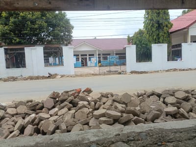 The image shows a street scene in Meunasah Raya, Indonesia, with a pile of broken paving stones in the foreground. In the background, there is a white fence with metal bars and a building with a red tiled roof, which appears to be a school or public institution. The building has signs with Indonesian text and blue and white colored walls. There are trees on either side of the building and utility lines crossing the sky. The area appears to be under construction or undergoing renovation, as indicated by the pile of debris and building materials.