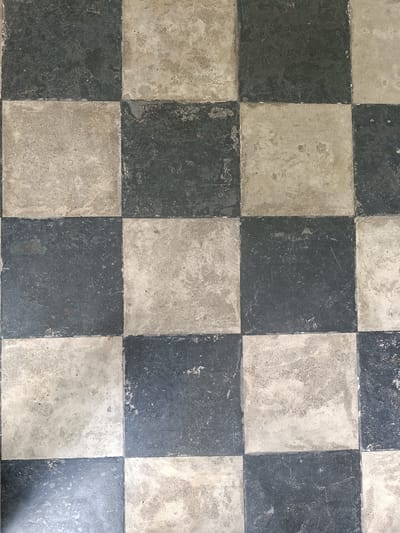 This image displays a close-up view of a weathered, black and white checkered floor. The tiles appear to be made of stone or concrete and show signs of age, with scuff marks, chips, and variations in color. The pattern is a classic checkerboard, with alternating dark and light squares. The lighting suggests it could be daytime, but no specific time can be determined. There are no people or objects visible in the frame, and no text is present. The setting is an interior space, likely a room with a tiled floor. The overall impression is one of an old, possibly historic, but well-used surface. The location context provided is Краснабярэжскі сельскі Савет, Belarus.