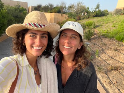 This image, captured in Aljezur, Portugal, depicts two adult females posing closely together in an outdoor landscaped setting. The individual on the left has dark, curly hair and is wearing a straw cowboy hat with a red, green, and white striped band. She has gold hoop earrings and is dressed in a light-colored, vertically striped button-down shirt. A light-colored towel is draped over her left shoulder, and a brown bag strap is visible. She wears multiple necklaces, including one with a blue, eye-shaped pendant. She is smiling broadly with visible teeth.

The individual on the right has dark, wavy hair and is wearing a light-colored baseball cap with the word "NIHI" visible on the front. She is dressed in a dark, possibly charcoal or black, long-sleeved shirt and wears multiple delicate gold necklaces. She is also smiling, facing the camera.

The environment features a grey paved pathway composed of interlocking pavers visible behind the left individual. To the right, a terraced garden area with dry earth, various green plants, and black drip irrigation lines is prominent. In the background, a light-brown, flat-roofed building structure is partially visible, featuring small, recessed rectangular details. Beyond the immediate landscape, lush green vegetation and trees are present on elevated ground, with a clear sky overhead. Two distant figures are discernible on a higher grassy slope in the far background. The scene is illuminated by natural sunlight, indicating daytime.