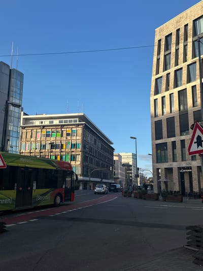 The image depicts a street scene in Osnabrück, Germany. The sky is clear and blue. Buildings flank the street. One building is a modern structure with many windows arranged in a grid pattern, and "L'OSTERIA" signage is visible. A road sign with a white arrow pointing left is near this building. Another building is also visible, with many colorful square panels or windows. A green "MetroBus" labeled "[Strom!] fahren!" is partially visible on the right side of the image, suggesting it is driving along the road. Several cars and pedestrians can be seen further down the street. The road is asphalt, with a designated red bus lane along the side. The image is oriented vertically, but the scene is rotated about 90 degrees.