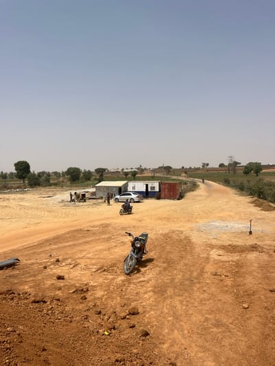This image depicts a dry, rural or semi-urban landscape in Gyellesu, Nigeria, under a hazy, light blue sky. The foreground consists of reddish-brown dirt and scattered debris, with a silver motorcycle parked facing slightly left. In the midground, a dirt road curves from the left towards the right, leading into the distance. To the left of the road, a small group of approximately three individuals stands near a cement mixer, actively engaged in a task. A light-colored sedan is parked nearby, next to a corrugated metal structure and two modified shipping containers, one blue and white, and another red. A person rides a dark motorcycle away from the viewer on the dirt road. Further along the road to the right, a shovel is stuck upright in the ground. The background reveals sparse green vegetation, scattered distant structures, and utility poles under a wide, open sky.