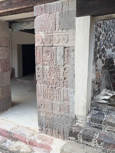 The image shows a close-up of stone carvings in San Francisco Mazapa, Mexico. The carvings are on rectangular stone blocks that form part of an architectural structure. The stone is a light reddish-gray and the carvings consist of geometric patterns and stylized depictions of objects, including spirals and other shapes. The background includes a rough stone wall, a smooth light-colored wall, and a dark opening suggesting a passageway or doorway. The overall impression is that of ancient or historical stonework.