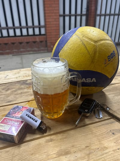 In Dinskaya, Russia, an image captures a still life arrangement on a wooden surface. The main subjects are a worn, yellow and blue volleyball and a glass mug filled with beer, showcasing a frothy head. Nearby, a set of car keys with a leather strap and a metal charm are placed, along with a white lighter branded "THE TURNER" resting on a pack of "CHAPMAN" cigarettes. The scene is illuminated by artificial light, suggesting it might be evening or indoors. The background shows a blurry industrial-like structure with horizontal slats and brickwork, indicating an outdoor or semi-outdoor setting. The presence of beer and cigarettes, along with the car keys, suggests a casual, possibly end-of-day, relaxation scenario. Visible text includes brand names like "MIASA," "CHAPMAN," and "TURNER," as well as Russian text on the cigarette pack, which appears to be a warning about health consequences, and a phone number. The overall mood is relaxed and unpretentious.